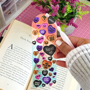 May include: A clear, arched bookmark featuring a colourful pattern of heart-shaped sweets with Halloween-themed text including Haunt Me, Spooky Season, Bite Me, and Trick or Treat, finished with a white tassel attached to the top centre.