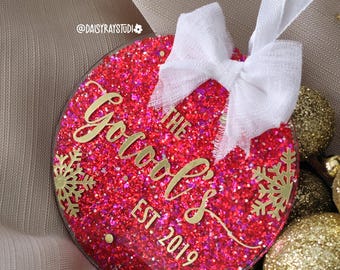 May include: A round, red glitter Christmas ornament with gold text reading "The Goods EST. 2019". It features gold snowflake designs and a white ribbon. The ornament is part of a festive holiday theme.