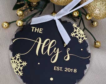 May include: A black and gold Christmas bauble with the text "The Ally's EST. 2018" and snowflake designs. The bauble is round with a white ribbon for hanging. Gold Christmas decorations and greenery are in the background.