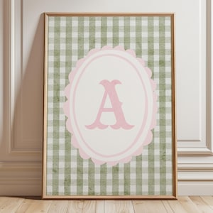 May include: A framed print with a pink letter "A" in a scalloped oval, on a green and white gingham background. The frame is light brown. The print is against a white wall and wooden floor. The overall design is a classic style.