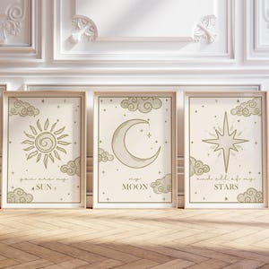 May include: Three framed art prints with celestial designs. The prints feature a sun, moon, and star, each with accompanying text. The color palette is neutral with beige backgrounds and light green illustrations. The frames are light wood.