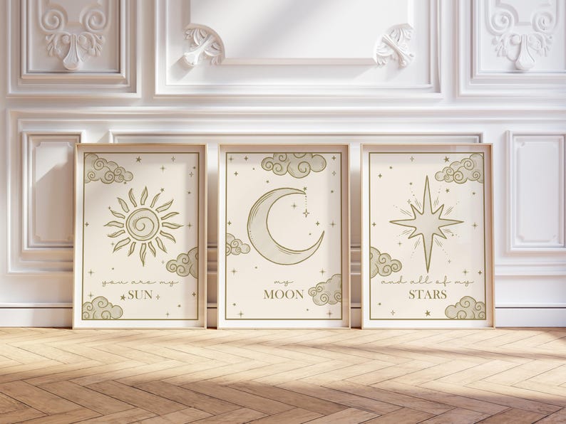 May include: Three framed art prints with celestial designs. The prints feature a sun, moon, and star, each with accompanying text. The color palette is neutral with beige backgrounds and light green illustrations. The frames are light wood.