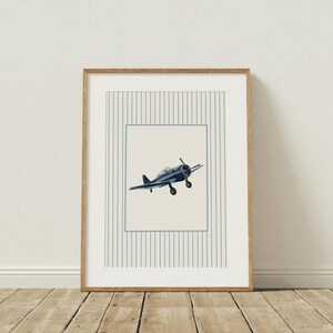 K&ouml;nnte beinhalten: Ein gerahmter Druck mit einer Illustration eines Marineblauen Vintage-Flugzeugs. Das Kunstwerk befindet sich vor einem cremefarbenen Hintergrund innerhalb eines blau linierten Rahmens und eines Streifenmusters. Der Rahmen besteht aus hellem Holz.
