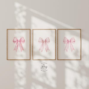 May include: Three vertical rectangular wall art prints in thin wooden frames, each featuring a centred watercolour illustration of a soft pink ribbon bow. Below the frames, the text reads All Things Rosy Art Collection in a circular logo.