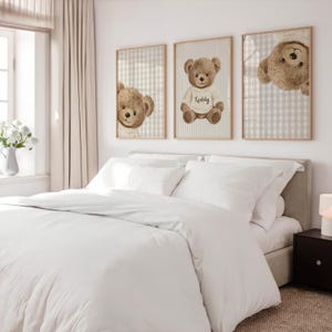 May include: Three framed prints of teddy bears above a bed with white bedding. The teddy bears are brown with cream-coloured shirts. One print shows a teddy bear wearing a shirt that says "Teddy". The frames have a light wood finish.