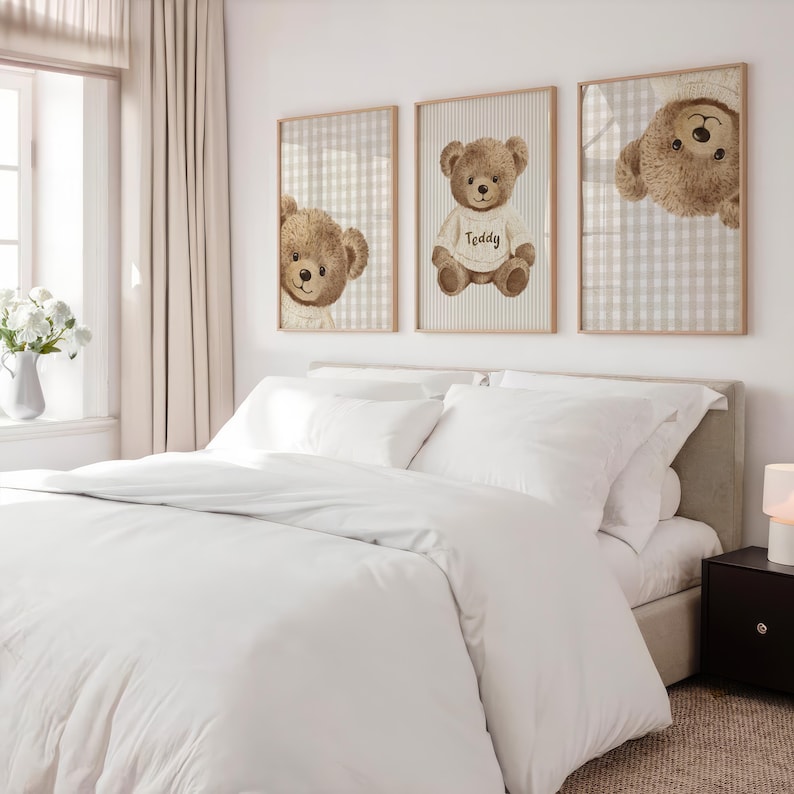 May include: Three framed prints of teddy bears above a bed with white bedding. The teddy bears are brown with cream-coloured shirts. One print shows a teddy bear wearing a shirt that says "Teddy". The frames have a light wood finish.