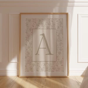 Pu&ograve; includere: Una stampa incorniciata con la lettera "A" in un colore tenue e neutro. La lettera &egrave; centrata all'interno di un bordo rettangolare, su uno sfondo a motivi floreali. La cornice &egrave; in legno chiaro e l'opera &egrave; appoggiata a una parete bianca.