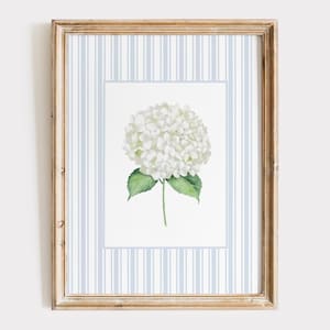 May include: A framed floral print featuring a white hydrangea bloom with green leaves and stem. The artwork is set against a light blue and white striped background, enclosed in a rustic wooden frame. The overall aesthetic is soft and elegant.