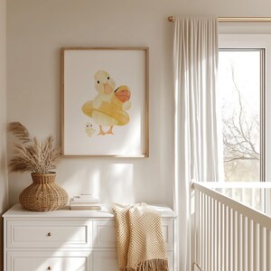 May include: A framed watercolor print featuring a mother duck and two ducklings. The ducks are yellow and orange, with the mother duck wearing a yellow inner tube. The print is displayed on a white wall above a white dresser.