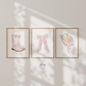 K&ouml;nnte beinhalten: Drei gerahmte Aquarelldrucke. Einer zeigt rosa Cowboystiefel, ein weiterer eine rosa Schleife und der dritte einen beigefarbenen Cowboyhut mit einer rosa Schleife. Die Drucke sind in Holzrahmen auf einer neutralen Wand. Text: "All Things Rosy Art Collection".
