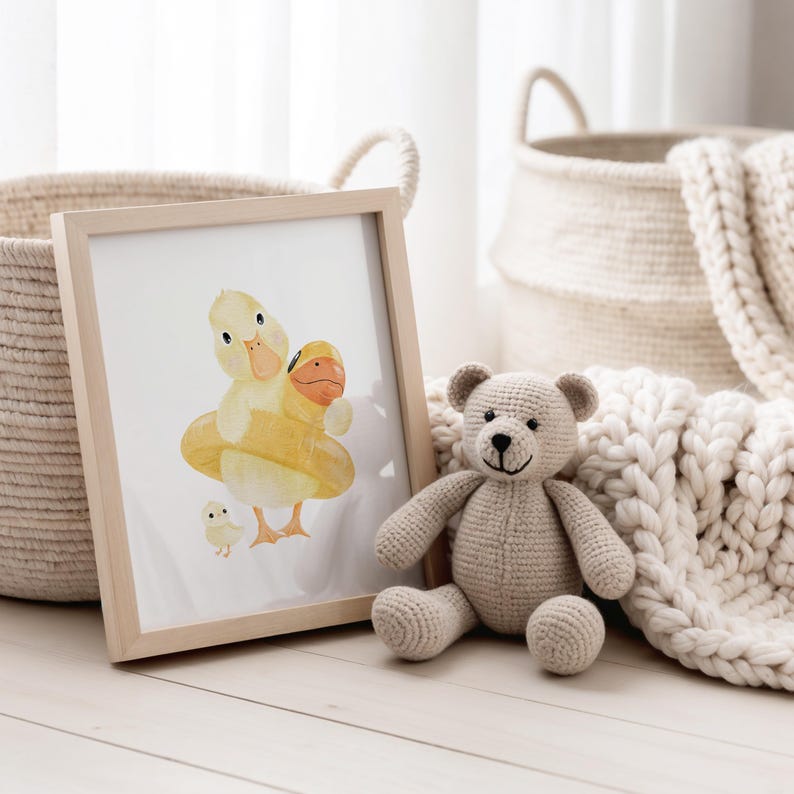 May include: A framed watercolor print of a yellow duck wearing a yellow inner tube, with a smaller duckling. A crocheted teddy bear sits next to the frame. Woven baskets and a cream-colored blanket are in the background.