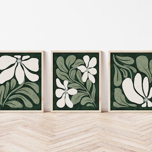 May include: A set of three framed floral art prints. Each print displays a white flower design with green leaves and stems against a dark green backdrop. The frames are light wood, and the art is on a wooden floor.