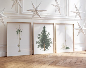 May include: Three framed Christmas art prints with gold frames, featuring a Christmas tree, bells, and a candle. White paper star ornaments hang from the ceiling. The prints are displayed on a wooden floor.