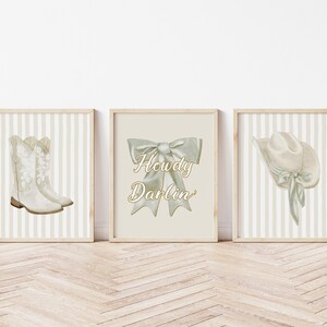 May include: Three framed prints with a western theme. The prints feature watercolor illustrations of cowboy boots, a bow with the text "Howdy Darlin'," and a cowboy hat. The backgrounds have a striped pattern in neutral tones.