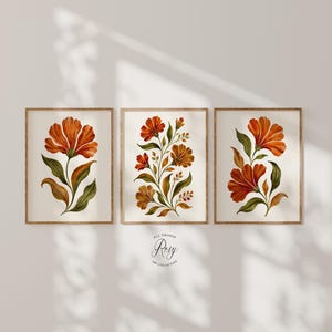 May include: Three framed wall art prints featuring botanical illustrations of orange flowers with green leaves on a cream background. The center print displays a cluster of flowers, while the side prints show single stems. Text reads All Things Rosy Art Collection.