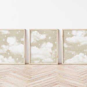 May include: Three framed prints showing a cloudscape with white clouds and stars on a beige backdrop. The frames are light wood, and the prints are displayed on a wooden floor. The overall theme is a dreamy aesthetic.