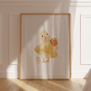 May include: Watercolor illustration of a mother duck and two ducklings. The mother duck is yellow with an orange beak and is holding a yellow floatie. The ducklings are yellow with orange beaks. The artwork is framed in a light wood frame.