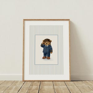 K&ouml;nnte beinhalten: Gerahmter Kunstdruck mit einem braunen Teddyb&auml;ren in einem blauen Fliegeranzug, braunen Stiefeln und einer Leder-Fliegerm&uuml;tze. Das Kunstwerk ist vor einem hellen Hintergrund mit vertikalen Streifen in einem Holzrahmen gerahmt.