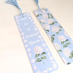May include: Two light blue bookmarks with blue tassels. One bookmark features the text "meet me at Cousins Beach" with seashell and hydrangea illustrations. The other bookmark has a hydrangea pattern.