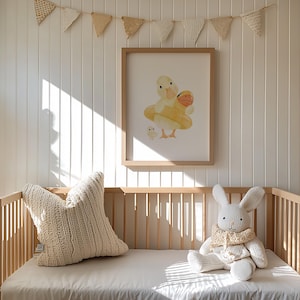 May include: A nursery scene featuring a framed watercolor print of a duck in a yellow floatie with a baby duck. A cream-colored knitted pillow and a stuffed bunny with a scarf sit on a crib with a white sheet. A banner of small flags hangs above.