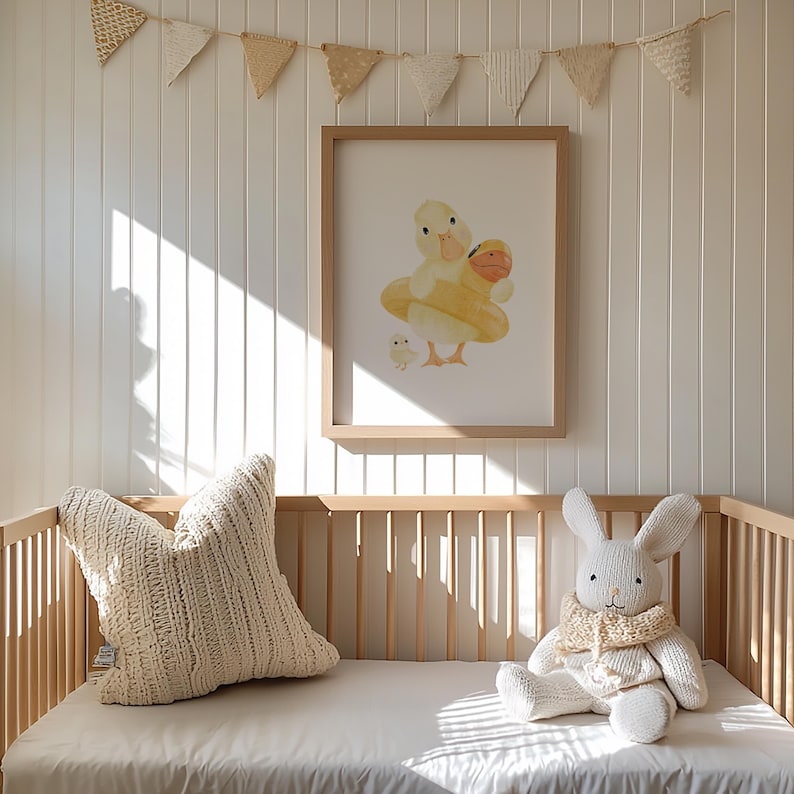 May include: A nursery scene featuring a framed watercolor print of a duck in a yellow floatie with a baby duck. A cream-colored knitted pillow and a stuffed bunny with a scarf sit on a crib with a white sheet. A banner of small flags hangs above.