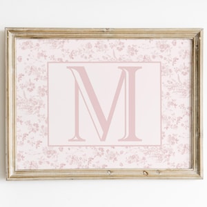 May include: A framed print with a large, pale pink letter "M" on a light pink background. The background has a floral pattern. The frame is a light brown colour, and the overall aesthetic is soft and feminine.
