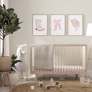 K&ouml;nnte beinhalten: Eine Szene im Kinderzimmer mit einem hellen Holzbett, rosa Bettw&auml;sche und einer grauen Decke. &Uuml;ber dem Bett befinden sich drei gerahmte Drucke mit rosa Illustrationen eines Cowboy-Stiefels, einer Schleife und eines Hutes. Ein Stofflama und eine Topfpflanze befinden sich ebenfalls im Raum.