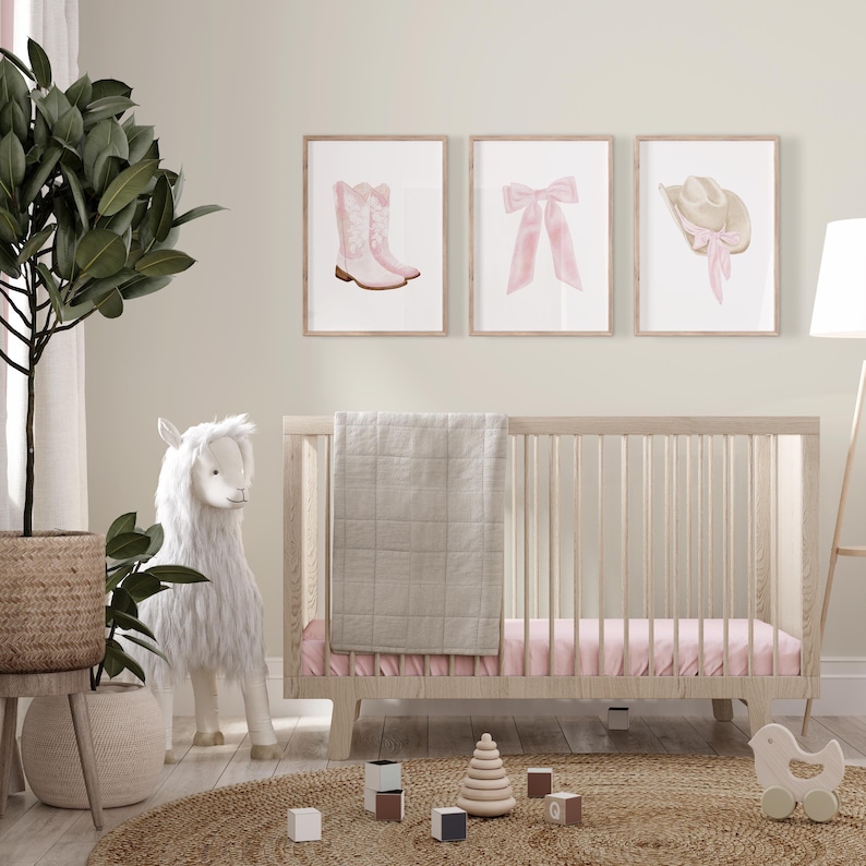 K&ouml;nnte beinhalten: Eine Szene im Kinderzimmer mit einem hellen Holzbett, rosa Bettw&auml;sche und einer grauen Decke. &Uuml;ber dem Bett befinden sich drei gerahmte Drucke mit rosa Illustrationen eines Cowboy-Stiefels, einer Schleife und eines Hutes. Ein Stofflama und eine Topfpflanze befinden sich ebenfalls im Raum.
