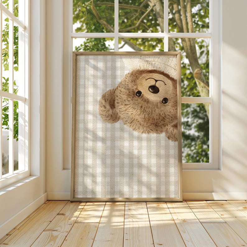 May include: A framed print of a teddy bear illustration. The bear is depicted in light brown with a smiling expression, set against a beige and white checkered pattern. The frame is light wood, and the print is displayed near a window, with natural light.
