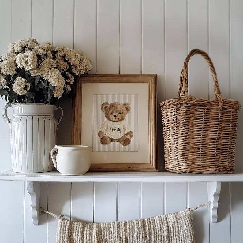 May include: A framed print of a teddy bear wearing a sweater with the name "Teddy" on it, displayed on a white shelf. A white ceramic vase with flowers, a small mug, and a woven basket are also on the shelf. The background is a white wall.