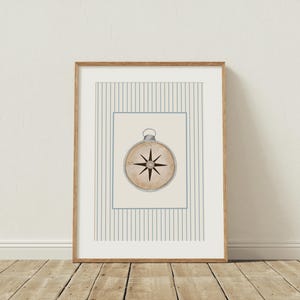 K&ouml;nnte beinhalten: Gerahmter Druck mit einer Vintage-Kompass-Illustration. Der Kompass befindet sich auf einem cremefarbenen Hintergrund, umrahmt von einem hellblauen Rand und einem gestreiften Muster. Der Rahmen ist aus hellbraunem Holz.
