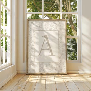 Pu&ograve; includere: Una stampa incorniciata con la lettera "A" in grigio chiaro, su uno sfondo chiaro con un motivo floreale. La cornice &egrave; color legno chiaro. La stampa &egrave; esposta in una stanza luminosa con pavimento in legno, vicino a una finestra.