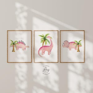 May include: Three framed watercolor prints featuring pink dinosaurs and palm trees. The dinosaurs include a Stegosaurus, a Brachiosaurus, and a Triceratops. The artwork has a soft, pastel color palette and a whimsical style, perfect for a nursery or children's room.