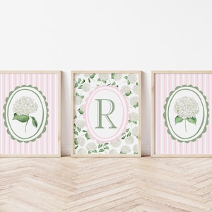 May include: Three framed floral prints. Two prints feature a white hydrangea in a green oval frame on a pink striped background. The center print has a floral background with a pink oval and a green letter "R".