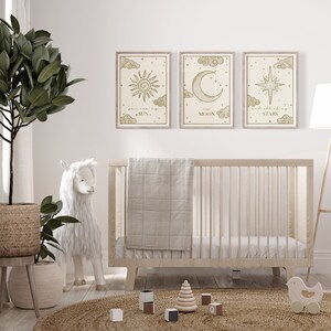 May include: Three framed art prints with celestial designs: a sun, a crescent moon, and a star, each with accompanying text. The prints are in a nursery setting, above a wooden crib with a neutral-colored blanket. A stuffed llama and a woven rug are also in the room.
