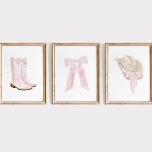 K&ouml;nnte beinhalten: Drei gerahmte Drucke mit Aquarellillustrationen. Die Drucke zeigen ein Paar rosa Cowboystiefel, eine rosa Schleife und einen Cowboyhut mit rosa Band. Die Rahmen haben eine hellbraune Holzoberfl&auml;che. Das Kunstwerk ist vor einem wei&szlig;en Hintergrund.