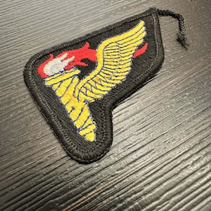 Puede incluir: Parche bordado negro con un ala amarilla y una antorcha con llamas rojas y blancas. El parche tiene un borde negro y un pequeño cordón negro adjunto en la parte superior. El diseño es un emblema estilizado.