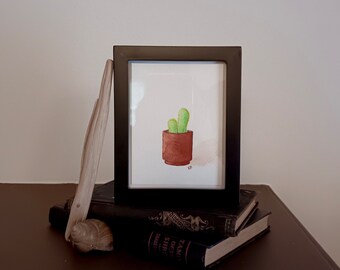 Peut inclure: Une peinture à l'aquarelle d'un cactus en pot dans un pot brun. La peinture est encadrée dans un cadre noir et repose sur une pile de livres.