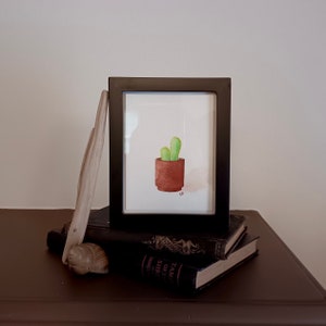 May include: A watercolor painting of a potted cactus in a brown pot. The painting is framed in a black frame and is sitting on top of a stack of books.