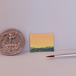 May include: A miniature painting of a sunset over a mountain range. The painting is on a small canvas and is about the size of a quarter. A small paintbrush is also in the image.