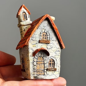 Puede incluir: Una casa de piedra en miniatura con tejado de tejas rojas y chimenea. La casa tiene pequeñas ventanas y una puerta de madera. La casa está sostenida en la mano de una persona. Es un objeto decorativo.