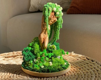 May include: A wooden sculpture of a woman with green moss hair and a moss base. The sculpture is on a woven wicker table.