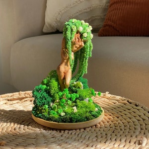 Puede incluir: Una escultura de madera de una mujer con cabello de musgo verde y una base de musgo. La escultura está sobre una mesa de mimbre tejido.