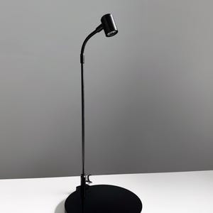 May include: A black desk lamp with a round base. The lamp has a flexible neck and a cylindrical light head. The base is approximately 8 inches in diameter. The lamp is on a white surface against a gray background.