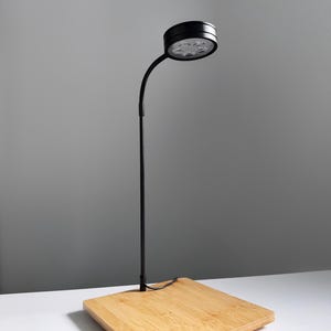 Puede incluir: Lámpara de escritorio LED negra con cuello de cisne flexible y cabezal de luz redondo. La lámpara está montada sobre una base cuadrada de bambú. Diseñada para iluminación enfocada.