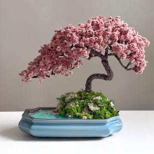 May include: A miniature bonsai tree with pink blossoms, a dark brown trunk, and green moss. The tree sits in a light blue ceramic pot with a water feature. The bonsai is a decorative piece.