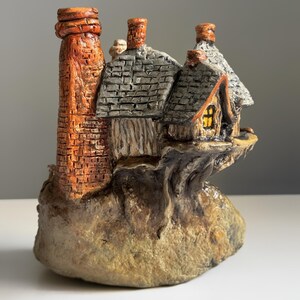 May include: A miniature cottage figurine with a brick chimney and a gray shingled roof. The cottage has a small window with a warm glow, and it sits on a rocky base. The color palette includes shades of brown, orange, and gray.