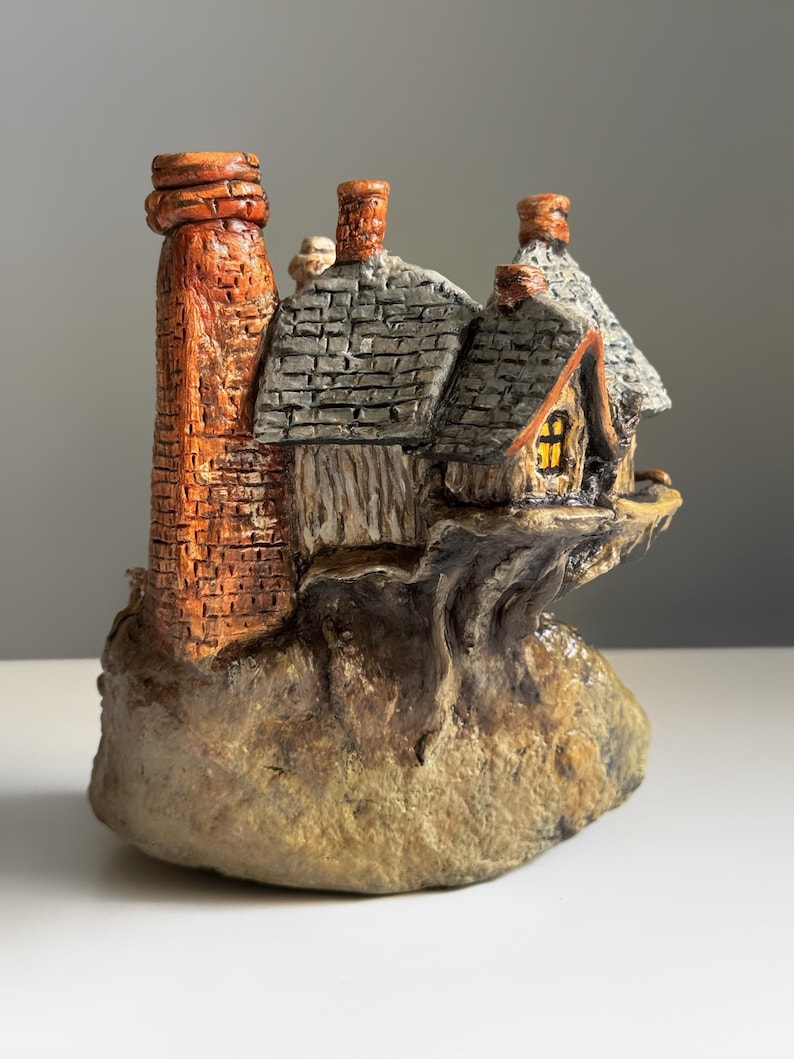 May include: A miniature cottage figurine with a brick chimney and a gray shingled roof. The cottage has a small window with a warm glow, and it sits on a rocky base. The color palette includes shades of brown, orange, and gray.
