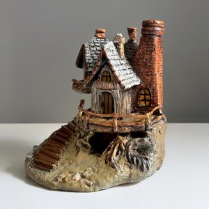 May include: A miniature ceramic cottage with a detailed design. The cottage features a brown wooden door, a chimney, and a stone base. The roof is white with a textured pattern. A small wooden staircase leads up to the cottage.