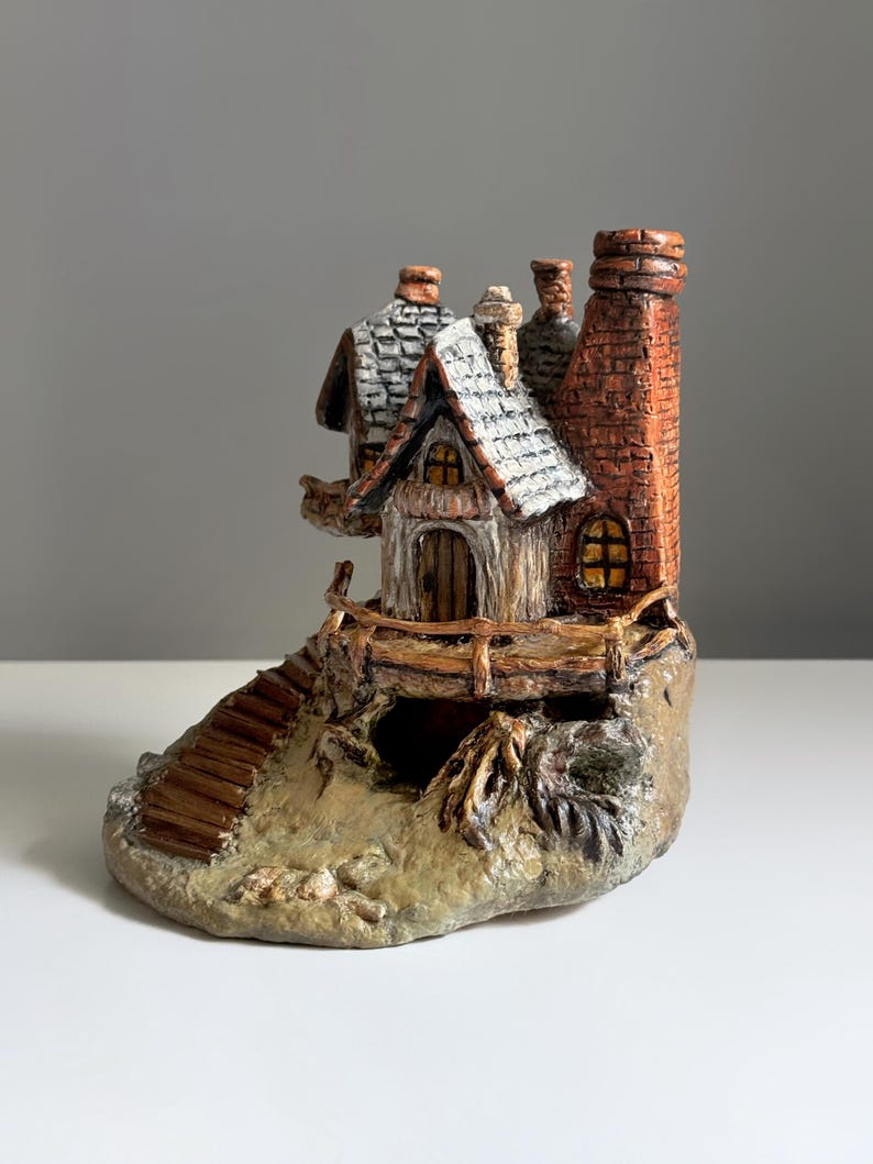 May include: A miniature ceramic cottage with a detailed design. The cottage features a brown wooden door, a chimney, and a stone base. The roof is white with a textured pattern. A small wooden staircase leads up to the cottage.